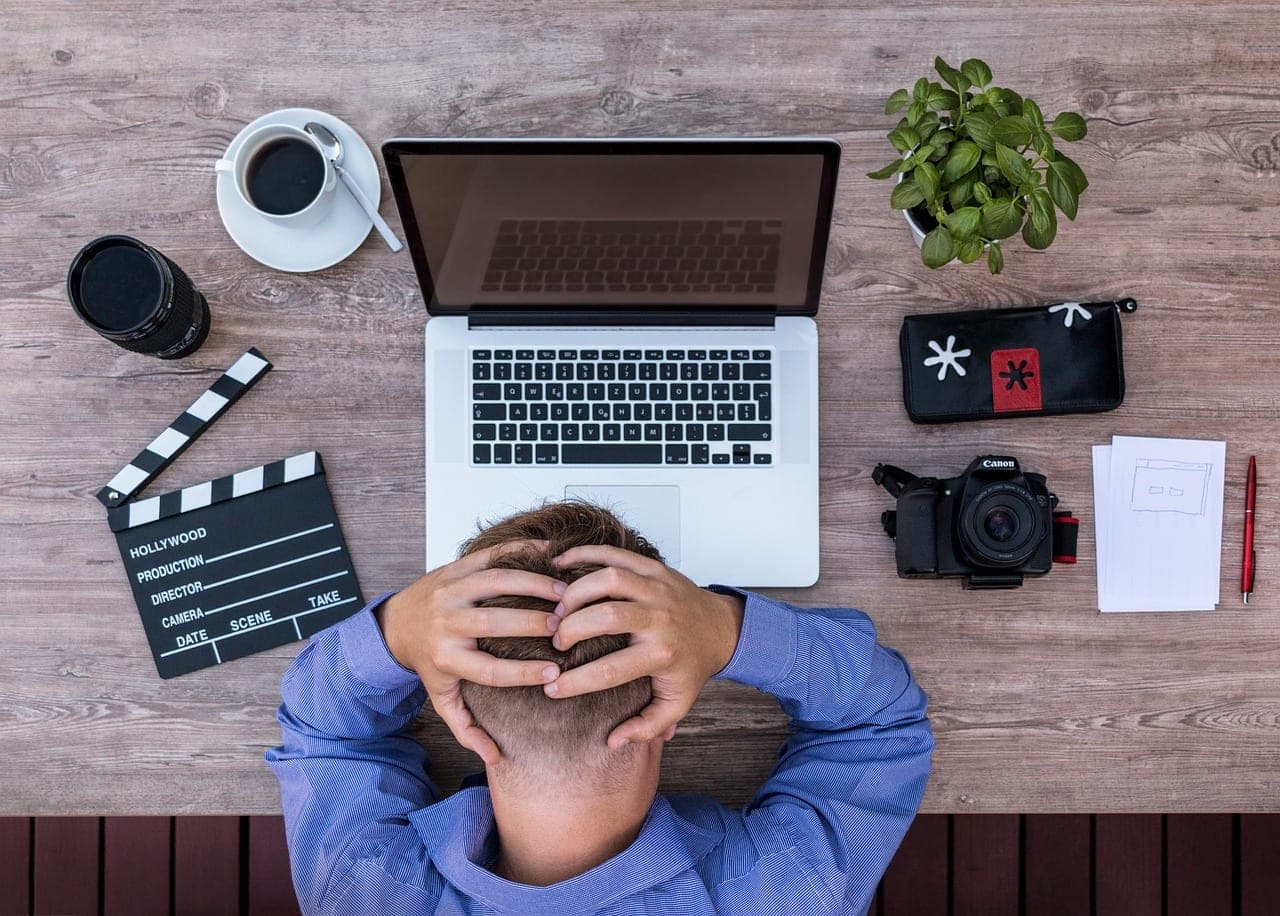 Vu d'en haut, un homme assis à son bureau, la tête dans les mains devant son ordinateur portable, entouré de carnets et d'une tasse
