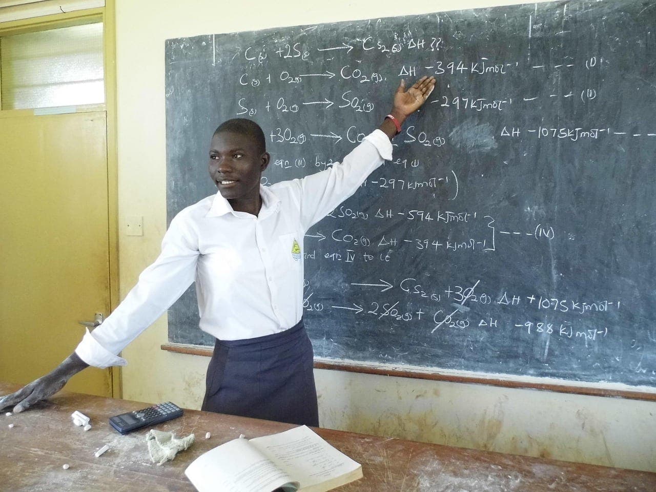 Un enseignant en chemise blanche, souriant, montre une formule de chimie écrite à la craie sur un tableau noir couvert d'équations