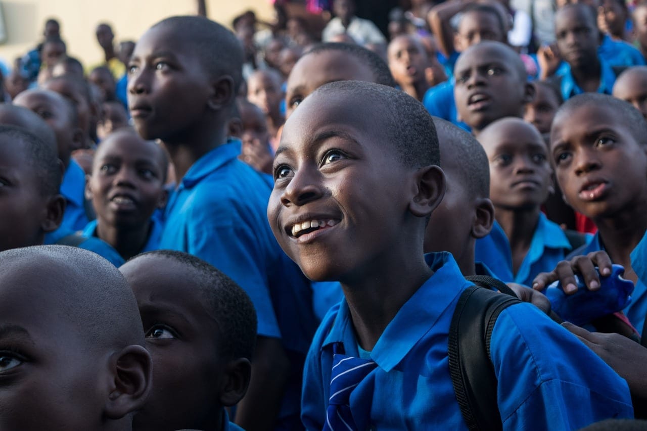 Des élèves en uniforme bleu, assis en groupe dans une cour d'école, écoutent attentivement ; un garçon sourit au premier plan
